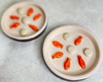May include: Two round ceramic plates with a cream-colored interior featuring a raised pattern of small orange fish and smooth cream-colored circular accents arranged in a circular design. The exterior of the plates is a dark brown color.