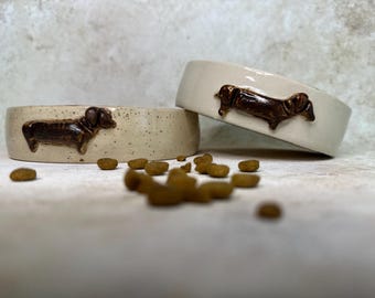 May include: Two ceramic pet bowls in a light cream color, each featuring a raised, dark brown dachshund figure attached to the outer side. The bowl on the left has a speckled finish, while the bowl on the right has a smooth, solid finish.