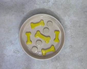 May include: A round, light beige pet bowl featuring a raised interior design with four bright yellow bone-shaped accents and three smooth, circular beige protrusions distributed across the base to create a textured feeding surface.