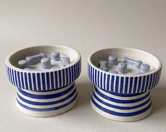 May include: Two ceramic slow feeder bowls featuring a white base with vertical and horizontal blue stripes. The interior of each bowl contains light blue raised obstacles in the shape of small bones and cylinders to slow down eating.