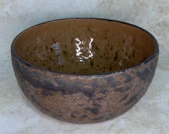 May include: A round ceramic bowl with a textured, mottled brown and dark charcoal exterior. The interior features a smooth, glossy tan finish with dark brown speckles throughout. The rim is uneven and dark, showing a handcrafted, organic appearance.