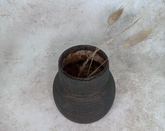 May include: A dark brown, textured ceramic vase with a rounded body and a wide, open neck. The vase features a rough, matte finish and contains three dried, light tan ornamental grasses extending from the opening, viewed from above.