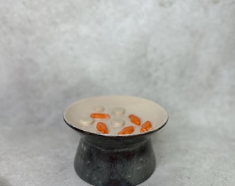 May include: A ceramic bowl with a wide, flared rim and a dark, mottled gray base. The interior of the bowl is a light cream color and features several small, raised, orange fish-shaped accents and circular bumps scattered across the bottom surface.