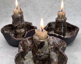 May include: Three dark brown, textured ceramic candle holders with irregular, wavy edges. Each holder features a central pillar containing a lit, off-white taper candle with visible wax drips pooling inside the bowl-shaped base.