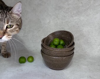 May include: A stack of three dark brown speckled ceramic bowls with a rough texture. The top bowl contains several small, round, bright green plums. Two additional green plums are positioned on the surface next to the stacked bowls.