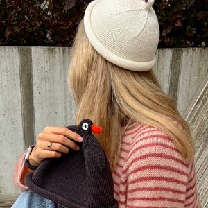 May include: A cream-coloured knit beanie with a small red and black chicken-shaped topper, paired with a dark charcoal knit beanie featuring a black, white, and red chicken-shaped detail, worn with a pink and white striped long-sleeve jumper.