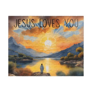 Puede incluir: Un rompecabezas que muestra un paisaje con un lago, montañas y un cielo al atardecer. Una figura está de pie en la orilla frente al agua. El texto JESUS LOVES YOU está impreso en negro en la parte superior. Las piezas forman una imagen completa.