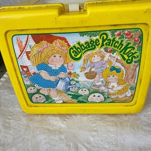 May include: Yellow plastic lunchbox with a Cabbage Patch Kids design. The lunchbox features a cartoon image of three Cabbage Patch Kids dolls in a garden setting. The text "Cabbage Patch Kids" is printed in a green and white font on the front of the lunchbox.