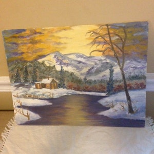 May include: An oil painting depicts a winter landscape with snow-covered mountains, a river, and a small cabin. The sky transitions from yellow to orange, and a bare tree stands on the right. The painting evokes a serene, natural scene.