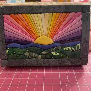 May include: A rectangular fabric art piece framed in dark gray fabric, featuring a sunburst design with rays in shades of orange, yellow, and pink. Below the yellow sun, a blue horizon line sits above rolling green hills with a leaf pattern.