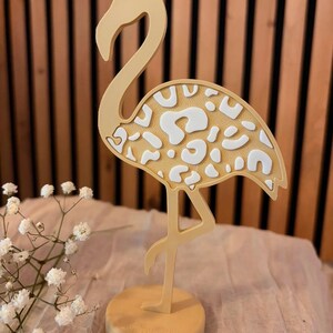 Könnte beinhalten: Eine stehende Flamingo-Figur in einem gedämpften Gelbton mit einer strukturierten Oberfläche am Körper und einem weißen Leopardenmuster. Der Flamingo ist auf einem runden Sockel montiert und steht auf einem Bein.