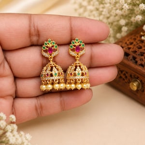 Puede incluir: Un par de pendientes jhumka dorados con un diseño en forma de campana adornado con pequeñas perlas blancas en la base, acentuados con piedras verdes y rosas incrustadas y cristales transparentes dispuestos en un patrón decorativo sobre el metal.