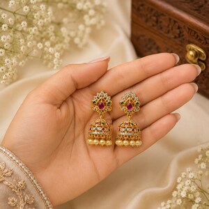 Pode incluir: Um par de brincos jhumka dourados com um design em forma de sino, adornado com pequenas pérolas brancas na base, cristais transparentes e pedras vermelhas centrais em forma de gota, acentuados com pequenas pedras verdes na estrutura.