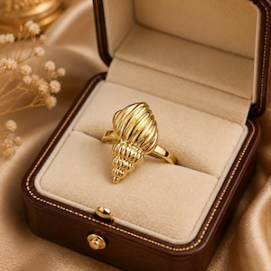 Può includere: Un anello dorato caratterizzato da un design a conchiglia lucido e testurizzato. L'anello è posizionato al centro di un portagioie rettangolare marrone scuro con coperchio incernierato e interno in velluto beige, che ne esalta la lucentezza.
