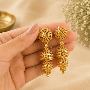 May include: A pair of gold-toned drop earrings featuring a circular stud top with a floral pattern, connected to two stacked bell-shaped jhumka pendants. Each component is decorated with intricate filigree and small bead-like textures throughout.
