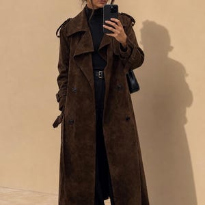 Pode incluir: Um sobretudo de camurça castanho-escuro com fecho de abotoamento duplo preto, lapelas largas, dragonas nos ombros e tiras com fivela nas mangas. É usado sobre uma blusa preta de gola alta, calças pretas com cinto e sapatos pretos de bico fino.