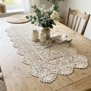 Könnte beinhalten: Ein cremefarbener, rechteckiger Häkel-Tischläufer mit gewelltem Rand und komplizierten Blumenmustern. Der Tischläufer liegt auf einem Holztisch mit zwei kleinen zylindrischen Kerzenhaltern und einer gefalteten beigen Leinen-Serviette.