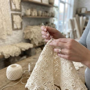 May include: An off-white crochet project featuring a repeating circular motif pattern. The textile is being worked with a thin metal crochet hook. A matching ball of off-white yarn sits on the wooden surface next to the unfinished lace piece.