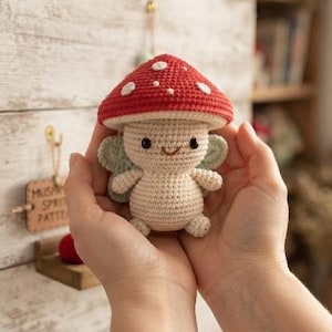 Könnte beinhalten: Eine kleine, handgefertigte Häkel-Amigurumi-Figur in Form einer Pilzfee. Die Figur hat einen leuchtend roten Hut mit weißen Punkten, einen cremefarbenen Körper und kleine hellgrüne Flügel. Sie besitzt aufgestickte schwarze Augen und ein Lächeln.