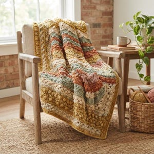 May include: A crochet blanket featuring a textured pattern with bobble stitches and stripes in shades of mustard yellow, sage green, terracotta, and cream. The blanket is draped over a light-colored wooden armchair, showcasing a complex, multi-layered design.