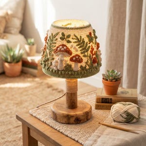Könnte beinhalten: Eine Tischlampe mit einem gehäkelten Lampenschirm, der einen cremefarbenen Hintergrund, grüne Farnblätter und orange-braune Pilze aufweist. Die Lampe hat einen zylindrischen Holzsockel, der mit Schnur umwickelt ist und eine natürliche Optik bietet.