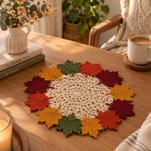 Könnte beinhalten: Ein rundes, cremefarbenes Häkeldeckchen mit einem dekorativen Rand aus kleinen, einzelnen Ahornblattmotiven in herbstlichen Farben, darunter Dunkelrot, Rostorange, Senfgelb und Waldgrün, die in einem kreisförmigen Muster angeordnet sind.