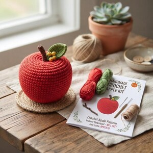 Könnte beinhalten: Ein roter gehäkelter Apfel mit braunem Stiel, grünem Blatt und kleinen gelben Beeren. Daneben liegt eine weiße Anleitung mit dem Titel HANDMADE CROCHET APPLE KIT, zusammen mit einem roten und einem grünen Garnknäuel sowie einer Häkelnadel.