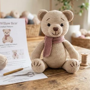 Könnte beinhalten: Ein beige gehäkelter Teddybär mit einem altrosa Schal, platziert neben einer gedruckten Anleitung mit dem Titel The Willow Bear, einem Knäuel beigem Garn, einer Holzhäkelnadel, einer kleinen Holzspule und einer silbernen Schere auf einer Holzfläche.