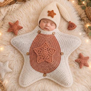 Puede incluir: Un saco de dormir para bebé tejido a ganchillo con forma de estrella grande en colores crema y terracota, que incluye un gorro puntiagudo a juego con una pequeña estrella, cuatro botones de madera y un motivo central de estrella.