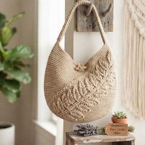 Könnte beinhalten: Eine beige Häkel-Hobo-Tasche mit einem markanten, erhabenen Farnblattmuster auf der Vorderseite, einem einzelnen Schulterriemen und einem Holzknebelverschluss. Ein kleines rechteckiges Schild trägt die Aufschrift "Handmade Fern Stitch Hobo".