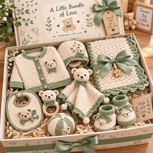 May include: A gift box set containing a cream and sage green crocheted cardigan, bonnet, blanket, bib, booties, a bear-themed rattle, and a plush toy. The box lid features the text A Little Bundle of Love and a bear illustration.