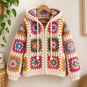 Op de afbeelding: Een hoodie met lange mouwen en ritssluiting, gemaakt van veelkleurige granny square gehaakte vierkantjes. Het kledingstuk heeft een crèmekleurige rand rond elk vierkant, een centrale metalen rits en twee lange crèmekleurige trekkoorden.