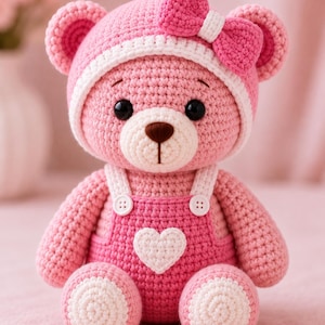 May include: A crocheted teddy bear in shades of pink and white. The bear wears a pink hat with a white trim and a pink bow, and pink overalls with white buttons and a white heart patch on the chest. It has black eyes and a brown stitched nose.