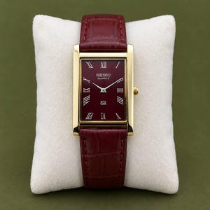 Op de afbeelding: Een rechthoekig Seiko Quartz horloge met een goudkleurige kast en een diepbordeauxrode wijzerplaat. Het horloge heeft Romeinse cijfers als uurmarkeringen, zilverkleurige wijzers en een bordeauxrode leren band met krokodillenprint.