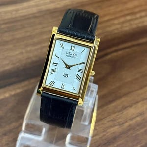 Può includere: Un orologio da polso rettangolare Seiko Quartz con cassa color oro e quadrante bianco con numeri romani neri. L'orologio presenta un motivo centrale testurizzato, lancette color oro e un cinturino in pelle nera con texture a coccodrillo.