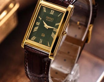 May include: A rectangular Seiko Quartz watch featuring a dark green dial with gold-tone Roman numeral hour markers and matching gold hands. The watch has a polished gold-tone case and a dark brown leather strap with a textured crocodile-style finish.