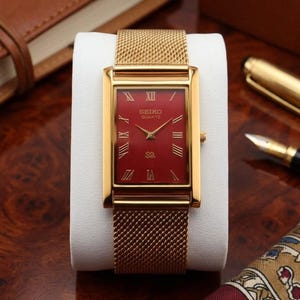 Op de afbeelding: Een rechthoekig goudkleurig polshorloge met een felrode wijzerplaat en gouden Romeinse cijfers als uurmarkeringen. Het horloge heeft een goudkleurige metalen mesh-band en toont de tekst Seiko Quartz op de wijzerplaat.
