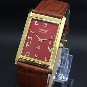 May include: A rectangular Seiko Quartz wristwatch featuring a gold-tone case, a textured red dial with gold Roman numeral hour markers, and a brown leather strap with a crocodile-embossed pattern. The watch face displays the Seiko Quartz logo.