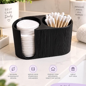 May include: A black, oval-shaped bathroom organiser with a ribbed exterior texture. It features two distinct compartments: one side holds a stack of round white cotton pads, while the other side contains a cluster of white cotton swabs with wooden sticks.