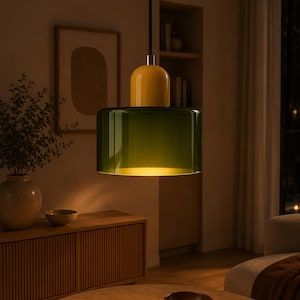 May include: A pendant light featuring a cylindrical, translucent dark green glass shade suspended from a black cord. The top of the fixture is a smooth, rounded yellow cap that connects to the glass body, emitting a warm, soft glow from within.