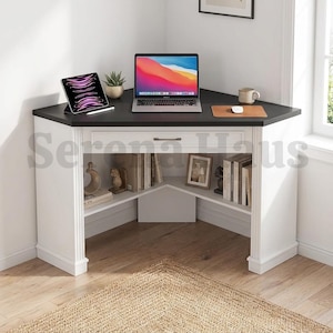 May include: A white corner desk with a black top, featuring a central drawer and open shelving on both sides. The desk surface holds a laptop, a tablet, and a mug on a mat. The shelves contain books and decorative items, with fluted side columns.