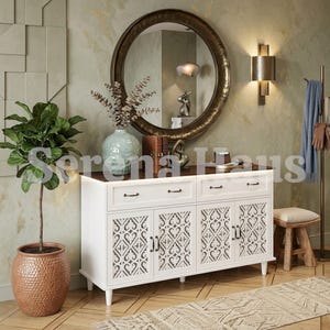 May include: A white wooden sideboard with two drawers and four cabinet doors featuring intricate cut-out patterns. The sideboard has a dark brown top surface and black metal hardware. It is positioned below a large round mirror with a bronze frame.