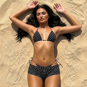 Könnte beinhalten: Ein schwarzer Bikini mit weißem Punktemuster. Das Oberteil ist im Triangel-Stil mit dünnen Trägern gehalten, während das Unterteil als tief sitzende Shorts mit seitlichen Bindebändern gestaltet ist. Die Badebekleidung besteht aus leichtem Stoff.