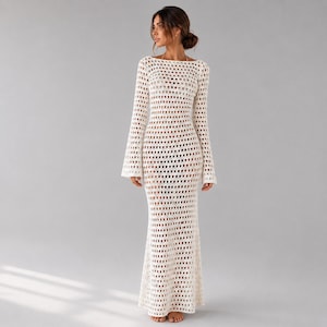 Puede incluir: Un vestido largo de ganchillo blanco de manga larga con un patrón de punto abierto en toda la prenda. El diseño presenta un cuello redondo y una silueta ajustada que se ensancha ligeramente hacia el dobladillo, con una puntada geométrica repetitiva.