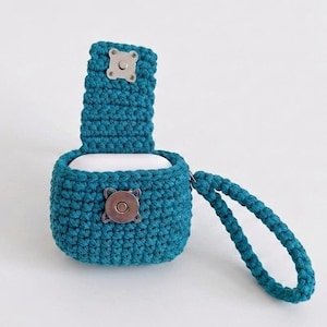 May include: A teal crochet case for wireless earbuds featuring a textured knit pattern, a vertical flap with a silver-toned magnetic snap closure, and a matching teal braided wrist strap attached to the side of the protective pouch.