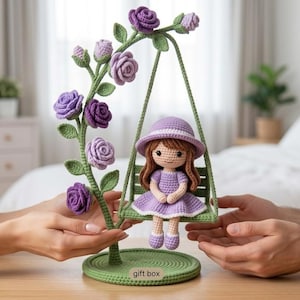 Puede incluir: Una muñeca de ganchillo con cabello castaño, vestido y sombrero morados, sentada en un columpio verde suspendido de un soporte curvo decorado con rosas moradas y hojas verdes. La base circular presenta el texto gift box en una etiqueta.