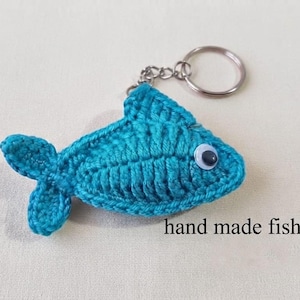 May include: A teal crochet fish keychain featuring a single plastic googly eye and a silver metal split ring. The fish is crafted with visible yarn stitches and includes the text hand made fish keychain printed in black in the lower right corner.