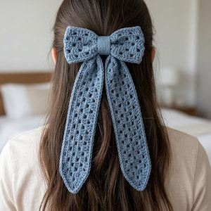 May include: A dusty blue crochet hair bow featuring a classic granny square stitch pattern. The accessory consists of a wide, rectangular bow section with a central knot and two long, dangling ribbon-like tails that extend downward from the center.