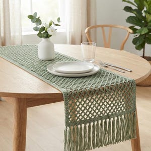 Puede incluir: Un camino de mesa de ganchillo en color verde salvia con un patrón de rombos abiertos y flecos largos en los extremos. El camino está colocado sobre una mesa de comedor redonda de madera, con un jarrón de cerámica y un plato blanco.