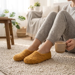 Puede incluir: Un par de zapatillas de ganchillo color amarillo mostaza con un patrón de punto texturizado y anudado. Las zapatillas son de estilo bajo y sin cordones, con puntera redondeada, diseñadas para la comodidad y el calor en interiores.